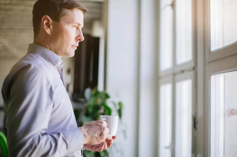 Mann steht mit Becher vor dem Fenster und guckt nach drau&szlig;en